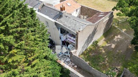 View from above of end terrace house - showing the front of the house missing due to explosion. The bunk beds are visible with debris scattered in front.