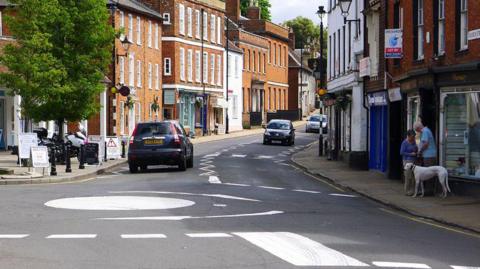 A small town street with brick buildings, two cars, a couple with a white dog near a shop, road markings including a large white roundabout.