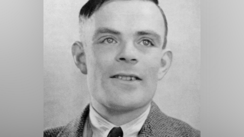 A black and white photo of a smiling Alan Turing - a man with short black hair and wearing a jacket, shirt and tie.