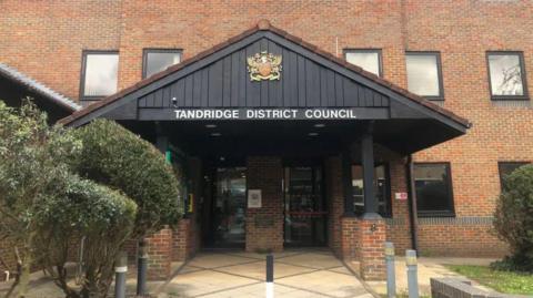 A triangular porch made from black wood, with the words TANDRIDGE DISTRICT COUNCIL across it in white and a coat of arms above it. The porch is on the front of a red brick building with half a dozen square windows on the front. To the left of the porch are a few small green hedges