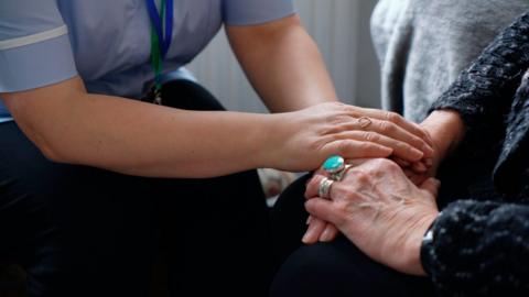 A female caucasian healthcare professional in purple uniform holds hands with an elderly caucasian woman who is wearing multiple silver and jade rings on her fingers and a dark knitted jumper. 