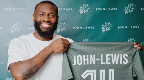 Footballer Lenell John-Lewis is wearing a white t-shirt and is smiling while holding up - at chest height - a dark green football shirt with thin stripes. The shirt displays the name “JOHN‑LEWIS” across the back and the number 14 in large white lettering. He stands in front of a green branded backdrop featuring repeated “John Lewis” text. 