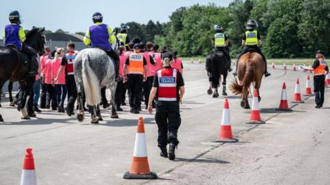 the backs of four officers on horseback as the horses walk behind and in front of a group of people, who can only be seen from the back. They are all wearing high visibility jackets
