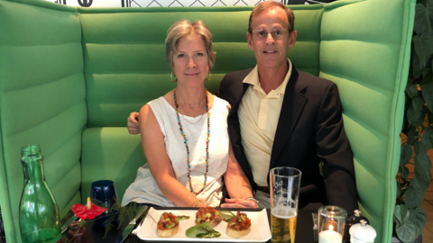 A couple sit smiling while sitting in a green booth at a table with food and drink on it