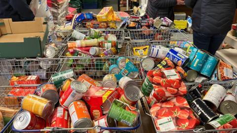 Approximately 10 standard sized shopping baskets full of tinned food such as tomatoes, baked beans and fruit sit on a long table.  The baskets also contain foodstuff such as pasta and crisps