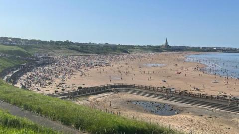 A view over Tynemouth beach with the sand packed with people. Some are walking around the empty old outdoor swimming pool in the foreground. Others are venturing into the sea. It is a sunny day with hundreds of people enjoying the sun.