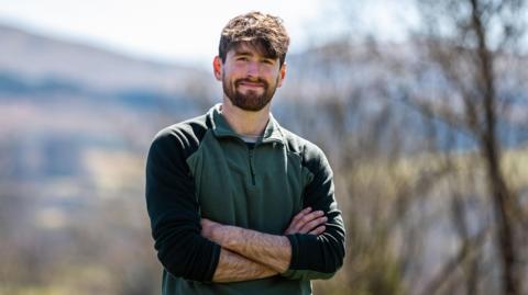 Rhys Evans, director of the Nature Friendly Farming Network in Wales, standing in a rural area, with his arms folded and smiling at the camera. He is wearing a green jumper with black on the arms. He has dark brown hair and a brown beard and moustache.