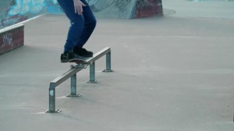 A skateboarder is riding on a pole in a skate park. He is wearing blue trousers and black shoes while riding the skateboard.