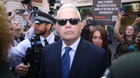 Huw Edwards, wearing a suit and sunglasses, outside court, surrounded by media and protestors