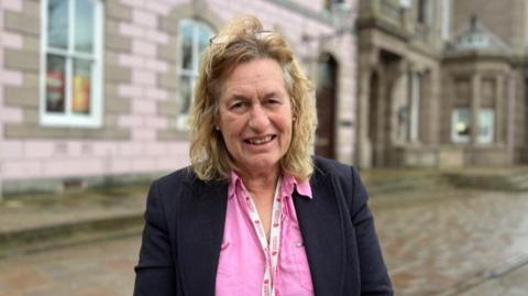 Andy Howell pictured in the Royal Square. The states assembly building is visible behind her. She is wearing a pink shirt and black jacket. She has blonde shoulder length wavy hair and a pair of reading glasses on her head. She is wearing a Government of Jersey lanyard. 