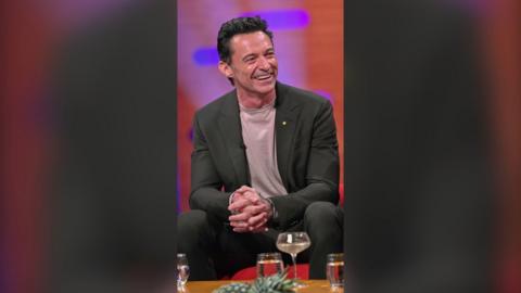 Hugh Jackman smiling in Graham Norton studio. He wears a grey suit with pale grey t-shirt underneath.