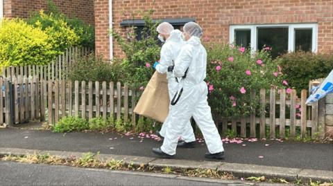 Two forensics officers in white suits are walking down a residential road.