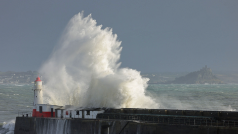 A wave hits a sea wall with a tower at the end.