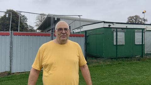 A man with a bald head and stubble is wearing glasses and an orange t-shirt. He is stood outside a construction side with fencing and a pavilion roof can be seen in the background