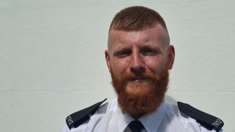 A man in a white and black police uniform standing in front of a blank, white wall.