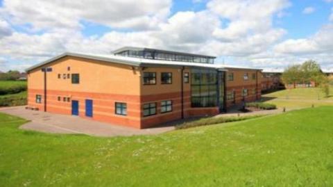 A modern two-storey L-shaped school building in the middle of a field. It has orangey-red brickwork which appears striped on the ground floor and there are blue doors. The sky above is blue with some white clouds.