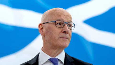 First Minister John Swinney standing in front of a Saltire backdrop