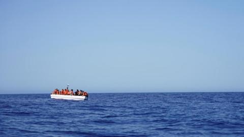 A vast open blue ocean and sky with a small white boat packed with people in orange lifevests