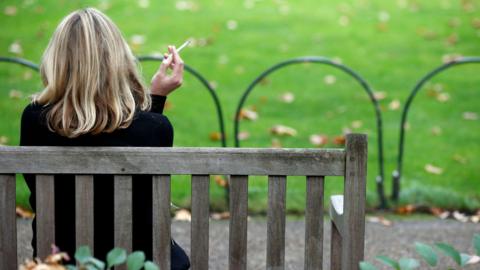 A woman with her back to the camera sitting on a park bench with a cigarette in her left hand. She has blonde hair just past her shoulders.