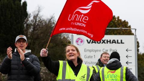 A woman in a high-vis jacket waves a red flag as workers protest outside the Vauxhall plant in Luton