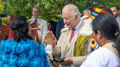 King Charles, in a suit and tie, meeting an Indigenous leader wearing a blue cloak made from feathers, at an event in Highgrove in July 2025.