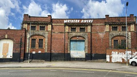 The exterior of the shed. It is built from red brick, has a number of boarded-up archways, mullioned windows and graffiti on the walls