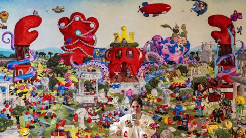 A man with brown hair wearing a white jacket and white-rimmed sunglasses holding his phone in front of him. He is standing with a wall-sized painting behind him featuring cartoon lobsters, octopus, buildings and emojis. 