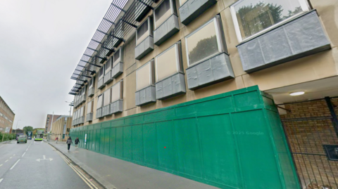 The picture is a streetview of Speedwell House in Oxford. It is a large building, stretching along the street, with protruding windows and green hoardings around the bottom floor.