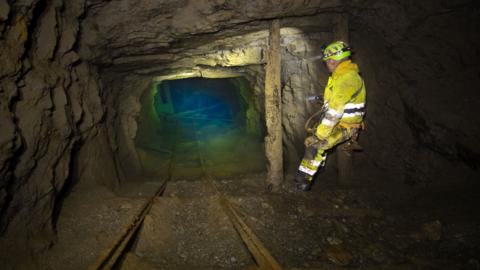 A man is a yellow boilersuit stands in an old mine working, the rocky tunnel ends in a green blue pool of water