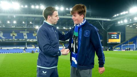 Seamus Coleman dressed in a blue tracksuit shakes hands with Mackenzie who is wearing a blue Everton top and scarf on the touchline at Goodison Park