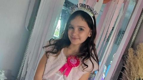 Aria smiles at the camera. She is wearing a pink birthday dress, a bright pink rosette and a birthday crown. She has long brown hair and the doorway behind her is decorated with pink streamers. 