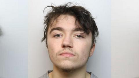 Mugshot of Kane Walker. He has some stubble, a moustache and brown hair. He looks expressionless. 