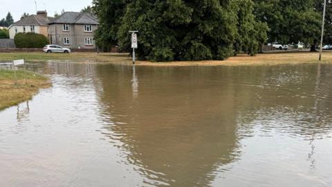 A road, where a junction would be, has flooded and the water has gone above pavement level. There are houses and trees in the background and there are several cars.