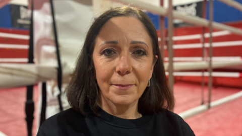 Kerry Norfolk was stood in the middle of the image looking towards the camera. She had shoulder length dark brown hair and was wearing a black jumper. She was stood in front of a boxing ring which had red floors and walls.
