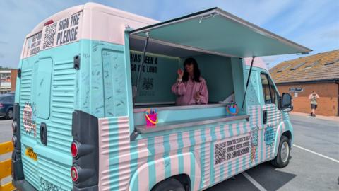 The ice cream van painted sky blue and pink parked in a car park the hatch is propped open and a person in a red striped shirt is inside