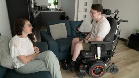 Jack in electric wheelchair next to his mother who is sitting on sofa in their living room