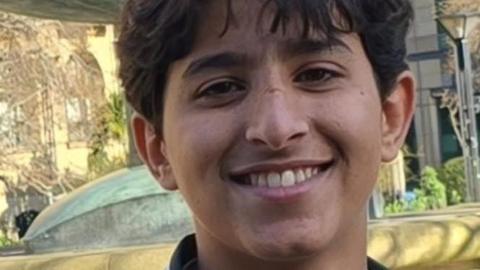 A boy smiles at the camera. He has brown hair, which falls over his forehead. The picture was taken outside, with several trees in the background.