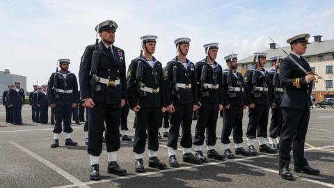 Royal Navy officers in uniform. There are srood in formation. The sky is blue with white clouds. 