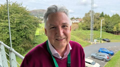Clive Summerfield stands on a balcony overlooking a car park. He is wearing a maroon jumper, pink shirt and green lanyard. He has grey hair and is smiling.