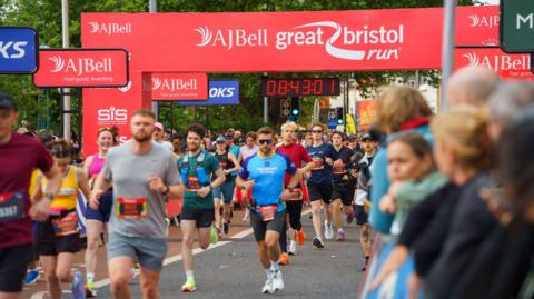 Dozens of people running down a road in Bristol. There is red canopy with the AJ Bell Great Bristol Run logo which has the race time on the clock.