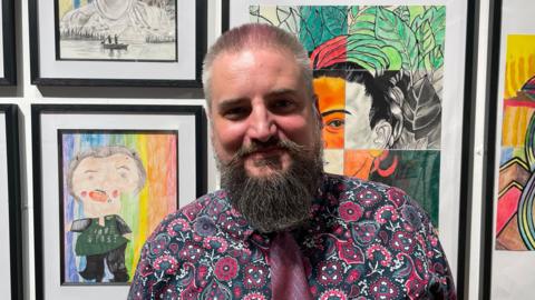 A man with short grey hair and a beard smiles into the camera. He is wearing a patterned pink and black shirt with a magenta tie and stands in front of a wall of self-portrait artwork.