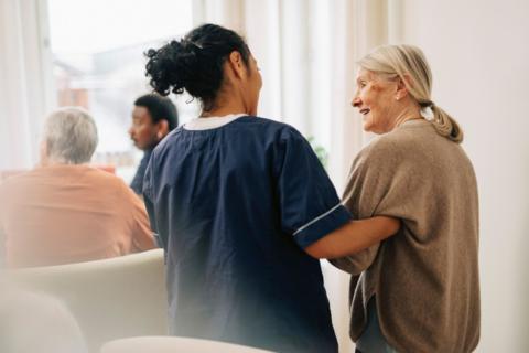 A woman in a nursing uniform has her back to the camera and is arm and arm with an older woman who has blonde hair in a pony tail. They are in a care home setting.