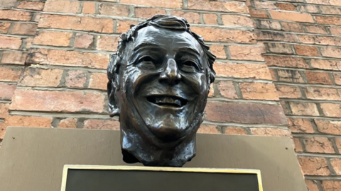 A bronze bust of a man's head with short hair in front of a brick wall