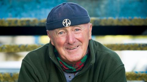 Dewi Pws Morris wearing his trademark skull-cap hat and badge, with a Welsh flag neck scarf, wearing a green fleece.