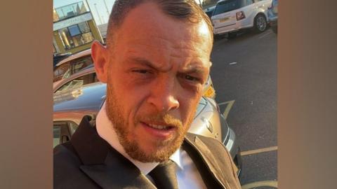 Aaron Heaney selfie. He is standing in a car park in the sun and wearing a white shirt, black tie and black suit jacket. He has short brown hair and a short beard