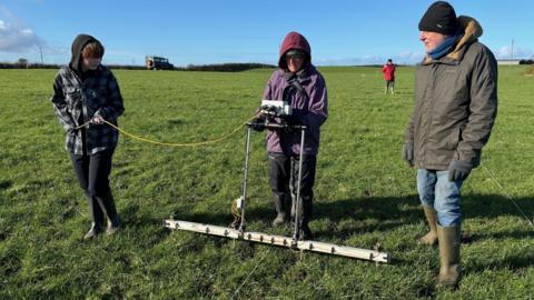 Three people standing in field holding a large stick-like structure