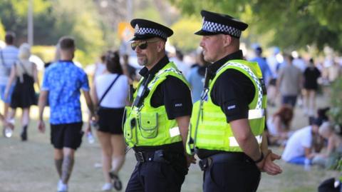 Two police officers, one wearing sun glasses, watch as Oasis fans walk by at Heaton Park