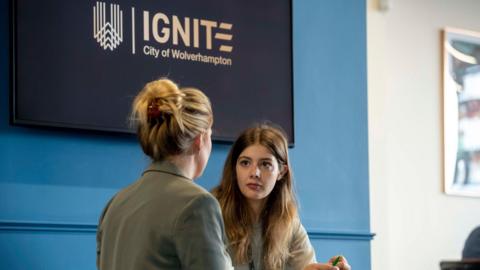 Two women are sat facing each other, of them them has their back to the camera. Behind them is a sign that reads, Ignite, City of Wolverhampton