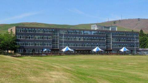 An old school building across four storeys set next to a playing field and with green rolling hills in the background