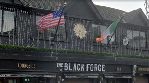 The sign of a pub that says The Black Forge in white writing on a black background. An American flag and an Irish tricolour are above. 